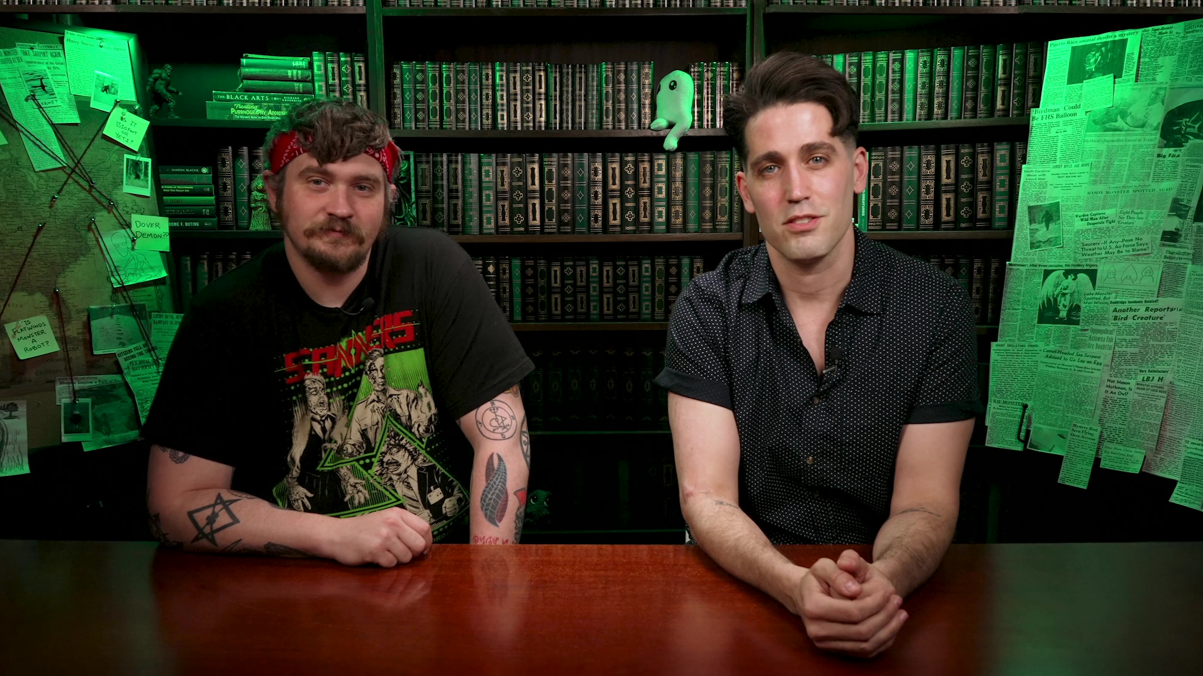 Peter Cross and Trevor Graham of Factor Entertainment are sitting at a desk in a studio with a bookcase and corkboards with newspaper clippings and an old map on the YouTube show, Cryptid Mysteries.